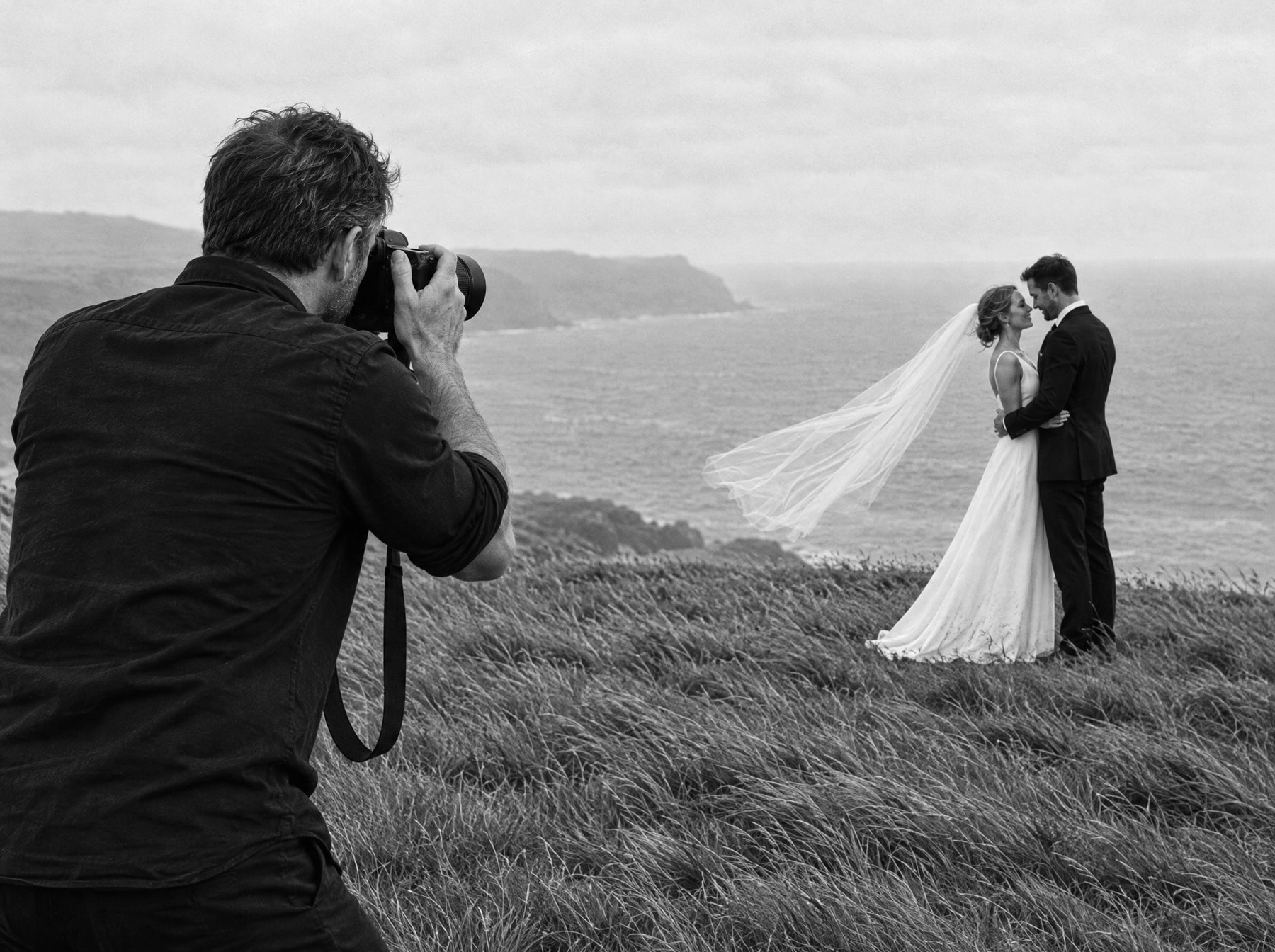 A wedding photographer, seen from behind, photographing a bride and groom standing together on a clifftop above the sea